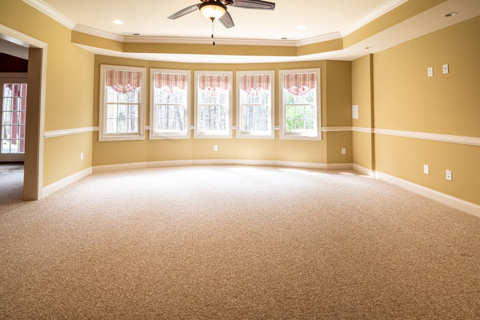 An empty living room with beige walls and cream-colored carpet flooring, featuring a large bay window with five sections, each adorned with pink and white striped valances. The room is well-lit by natural light streaming through the windows, with a ceiling fan centered in the ceiling. Brent Carpet Cleaning's expertise in surface cleaning and deep cleaning ensures this space remains hygienic, fresh, and spotless, highlighting the importance of regular sanitisation for a clean, inviting home environment.