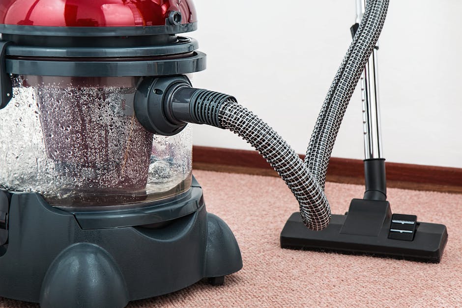 A woman using a yellow vacuum cleaner with a black hose attachment on a patterned, beige and pastel-colored area rug in a living room. The rug is placed on a wooden floor, with part of a red leather sofa visible on the right. She is dressed casually in a beige jacket, blue jeans, and white sneakers. The room is well-lit, and the scene depicts surface cleaning and deep cleaning practices by Brent Carpet Cleaning to maintain hygiene and freshness of the residential space.