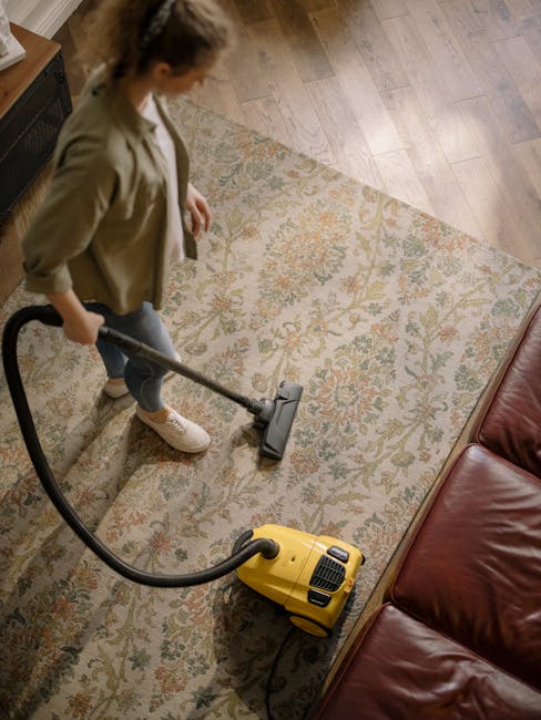A person operating a yellow vacuum cleaner on an ornate patterned rug in a living room, with wooden flooring nearby and a brown leather sofa partially visible. The individual is dressed casually and appears to be performing surface cleaning to remove dust and dirt from the carpet. Natural light illuminates the room, highlighting the cleanliness of the rug and surrounding area. This scene exemplifies professional deep cleaning, with a focus on maintaining hygiene and enhancing the appearance of domestic surfaces, as provided by Brent Carpet Cleaning, a specialist in cleaning services for residential and commercial spaces.