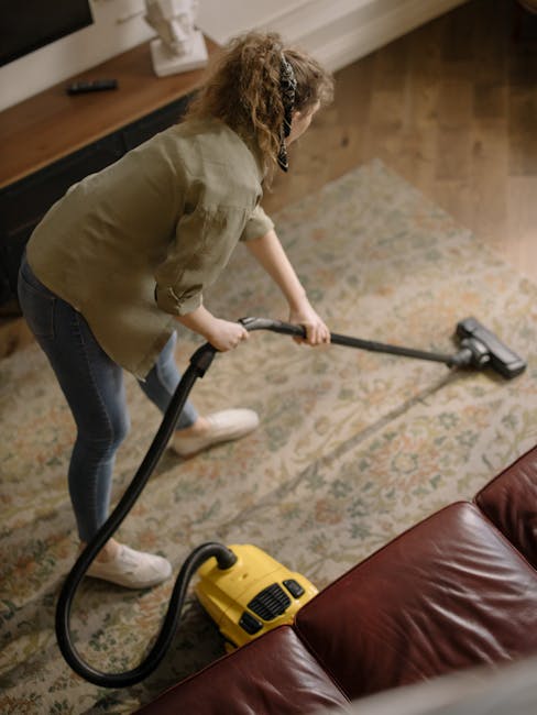 A woman with curly hair, dressed in a beige jacket, blue jeans, and white shoes, is performing surface cleaning using a yellow vacuum cleaner with a hose attachment on a patterned area rug in a living room. The room features wooden flooring adjacent to the rug, a low wooden cabinet with decorative items on top, and soft lighting illuminating the space. The rug appears clean and well-maintained, indicating a recent deep cleaning service provided by Brent Carpet Cleaning on Kilburn High Road. The scene emphasizes thorough domestic cleaning, including vacuuming and surface sanitisation, suitable for residential care in Brent.