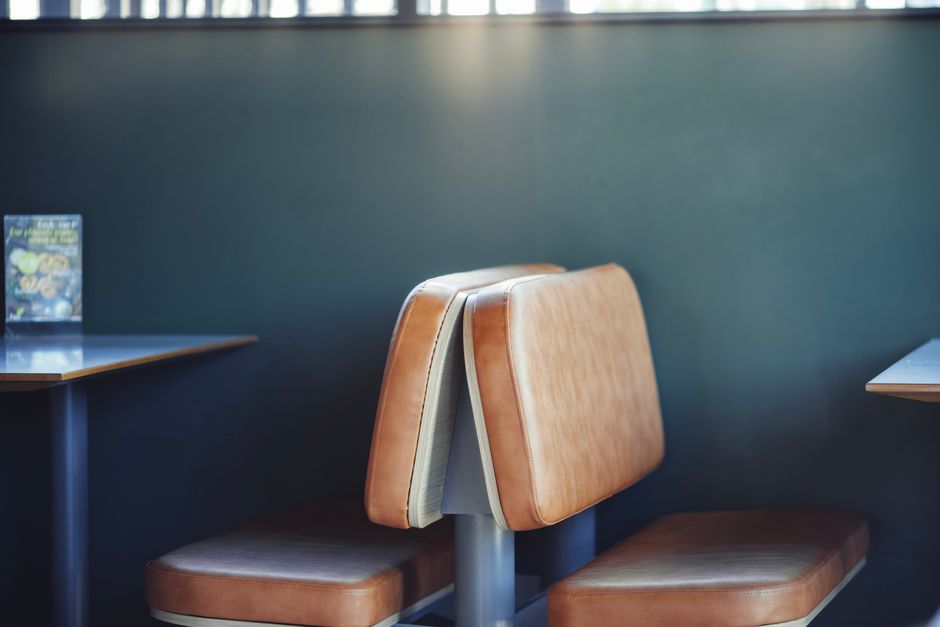 Interior view of a seating area featuring a pair of brown faux leather chairs with cushioned seats and backrests, positioned close together in front of a dark teal wall. To the left, part of a wooden table with a dark finish is visible, holding a small brochure or menu. The table and chairs are illuminated by soft, natural lighting, emphasizing the clean and tidy appearance of the furniture. The setting appears to be a commercial space, such as a café or waiting area, maintained by Brent Carpet Cleaning for effective surface cleaning and sanitisation of commercial environments, aligning with their same day upholstery cleaning services in Brent.