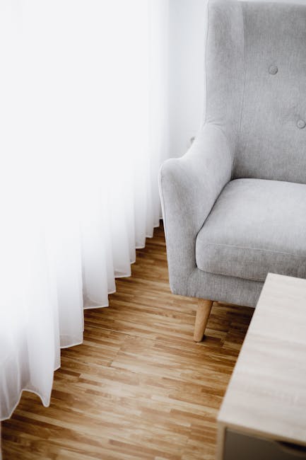 Image of a light grey upholstered armchair with wooden legs situated next to a window draped with white, semi-transparent curtains, allowing natural light to fill the room. The wooden floor beneath is clean and smooth, showcasing a natural finish. A small, light-colored wooden side table is placed nearby. The scene depicts a tidy, well-maintained living space, emphasizing cleanliness in furniture and flooring. Brent Carpet Cleaning offers surface cleaning and deep cleaning services aimed at maintaining hygienic, spotless domestic environments. The image highlights the importance of furniture and floor surface care as part of professional sanitisation routines.