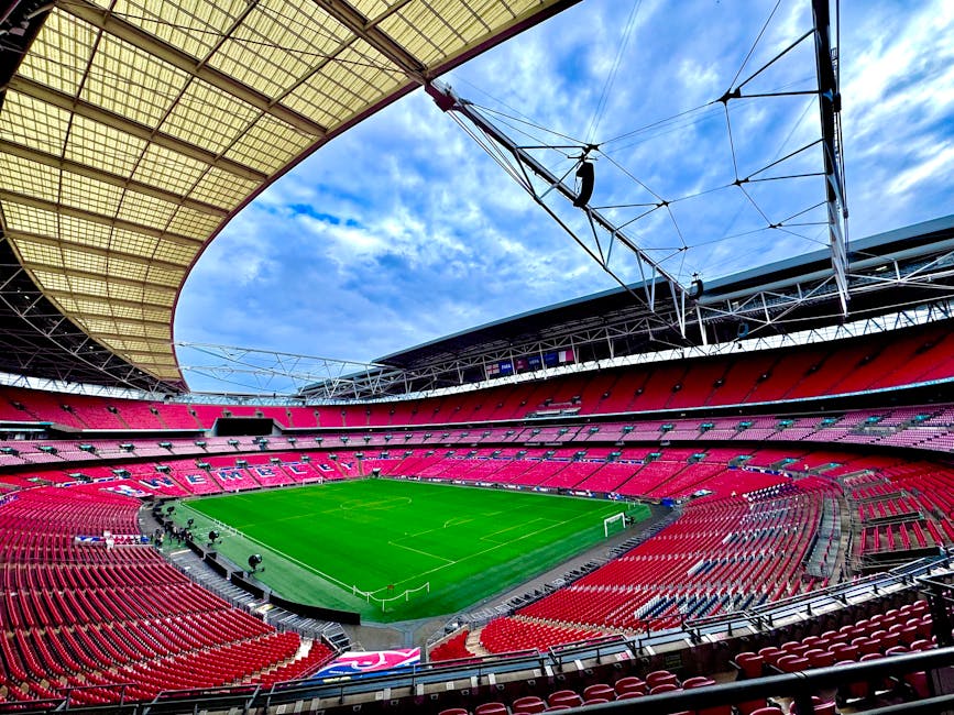 A wide-angle view of Wembley Stadium's interior showcasing a well-maintained, lush green football pitch surrounded by vibrant red and pink tiered seating. Overhead, a partly cloudy blue sky is visible through the stadium's roof structure, which includes a large, curved, semi-transparent canopy supported by steel cables. The stadium appears clean and tidy, with no visible debris or dust, reflecting thorough surface cleaning and maintenance by Brent Carpet Cleaning as part of their commercial cleaning services for large event venues.