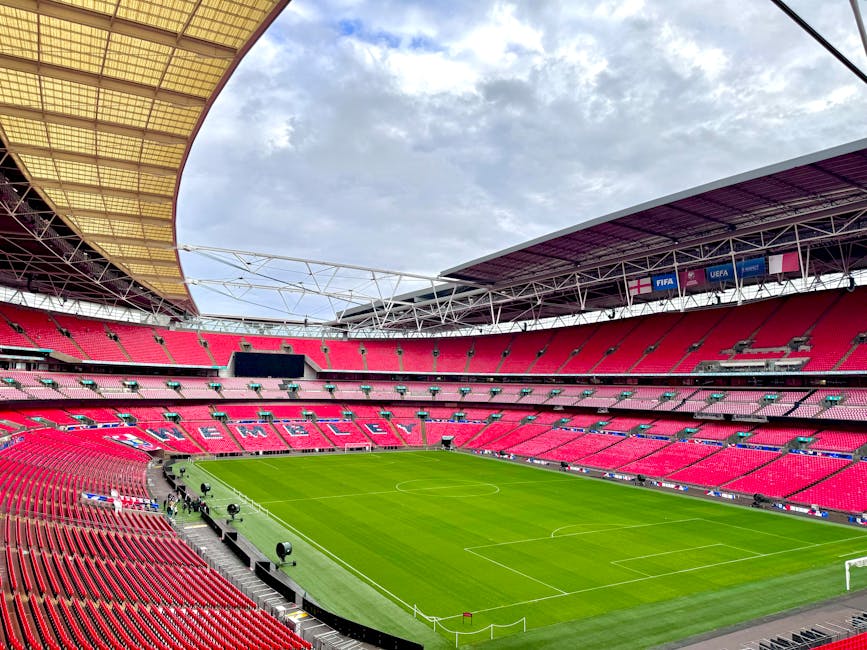 A panoramic view of Wembley Stadium's interior showing the well-maintained, lush green grass pitch with white boundary lines and goalposts at each end. Surrounding the field are rows of red stadium seats arranged in a curved pattern, with some areas shaded by the partial yellow and purple roof covering. The sky above features light clouds, and the stadium's modern infrastructure, including steel trusses and digital screens, is visible. The image emphasizes the clean and professional appearance of the sports venue, showcasing the thorough maintenance typical of Brent Carpet Cleaning's surface cleaning and deep cleaning services for large-scale facilities.