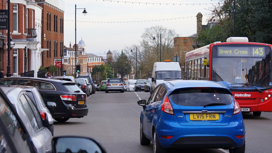 A street scene in Willesden Green showing parked cars along both sides of the road, including a blue hatchback with the license plate LV15 FRN in the foreground. A red Metroline bus with the destination sign 'Bren Cross 143' is passing by on the right. The street is lined with residential buildings featuring brick facades, white window frames, and some with small balconies. The road surface appears clean and well-maintained, with overhead streetlights and a cloudy sky overhead. The scene captures suburban urban infrastructure with a mix of parked vehicles, public transportation, and surrounding greenery, reflecting a typical residential area in Brent, London.