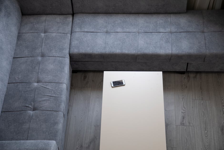 A top-down view of a grey fabric sofa with a sectional arrangement, situated in a well-lit living room with light wooden flooring. The sofa has visible stitching and a plush texture, appearing clean and well-maintained. In front of the sofa, there is a white rectangular coffee table with a smooth surface, on which rests a smartphone. The room's lighting highlights the cleanliness and fresh appearance of the furniture, with no dust or clutter visible. The image conveys professional surface cleaning and maintenance by Brent Carpet Cleaning, focusing on domestic cleaning and deep cleaning practices to ensure hygiene and surface freshness.